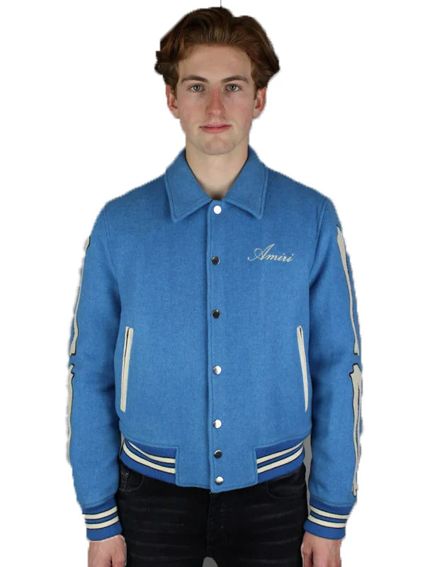 A young man with light brown hair models the Amiri Bones Jacket Mens by Zing Apparel, featuring a blue wool fabric, buttoned front, shirt-style collar, and "Amiri" embroidered on the chest, set against a white background.