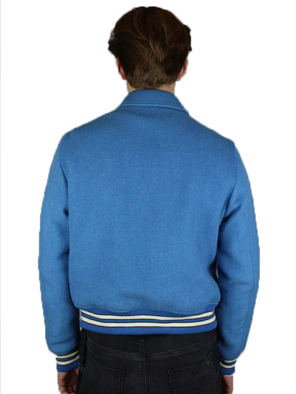 A person faces away from the camera wearing the Zing Apparel Amiri Bones Jacket Mens—a light blue wool jacket with buttoned front, white/navy striped cuffs and waistband—paired with dark pants against a plain white background.