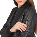 A woman with long brown hair unzips a small sleeve pocket on the front of the Zing Apparel Ava Ma-1 Leather Bomber Jacket for women in black, revealing part of her face.