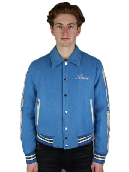 A young man with light brown hair models the Amiri Bones Jacket Mens by Zing Apparel, featuring a blue wool fabric, buttoned front, shirt-style collar, and "Amiri" embroidered on the chest, set against a white background.