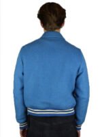 A person faces away from the camera wearing the Zing Apparel Amiri Bones Jacket Mens—a light blue wool jacket with buttoned front, white/navy striped cuffs and waistband—paired with dark pants against a plain white background.