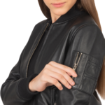 A woman with long brown hair unzips a small sleeve pocket on the front of the Zing Apparel Ava Ma-1 Leather Bomber Jacket for women in black, revealing part of her face.