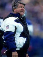 A football coach stands on the sideline during a game, wearing the Jimmy Johnson Cowboys Jacket by Zing Apparelâ€”a multicolor cotton-polyester NFL jacket with a hooded collar and a headset on, looking up.