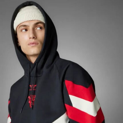 A person wearing a Zing Apparel Manchester United Originals Hoodie with red and white sleeve stripes and a white knit beanie looks off to the side against a plain gray background.
