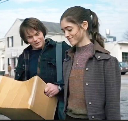 A young woman wears the Zing Apparel Stranger Things S01 Nancy Wheeler Purple Jacket while walking outdoors with a young man. Both are smiling, looking down at a large cardboard box, and chatting as they stroll along a street.