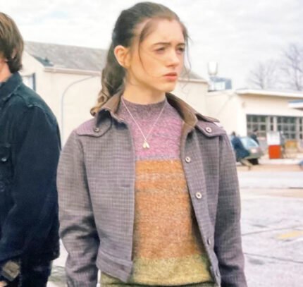 A young woman with long brown hair tied back, wearing the Zing Apparel Stranger Things S01 Nancy Wheeler Purple Jacket over a striped sweater and a necklace, stands outdoors in a parking lot with cars and buildings behind her.